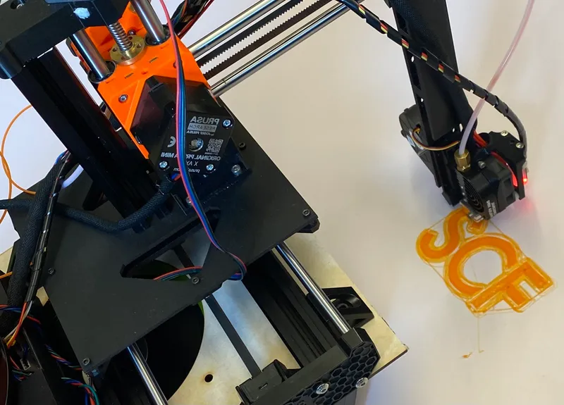 Photo of a cantilevered 3D printer as the print the letters "SCF" in orange plastic on a white floor surface.