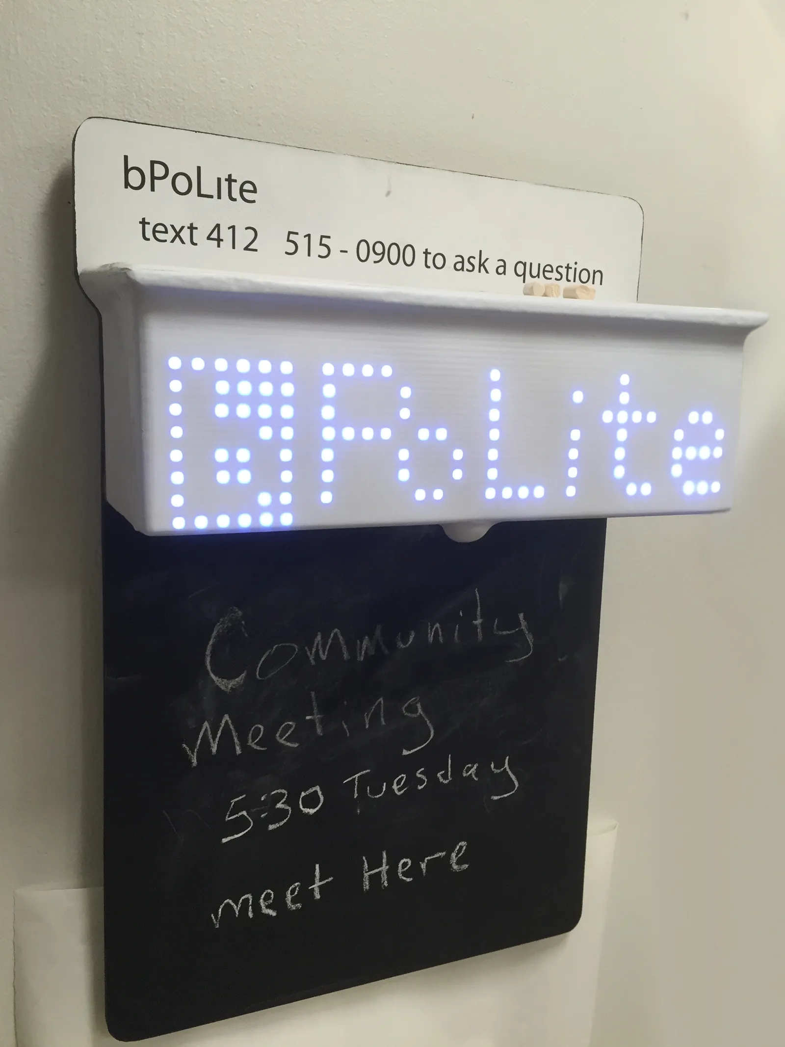 Photograph of the bPolite board with the name on top and a phone number encouraging users to text questions to the board. The board also has an LED display saying bPolite and a chalkboard attached to it where users can write using chalk.