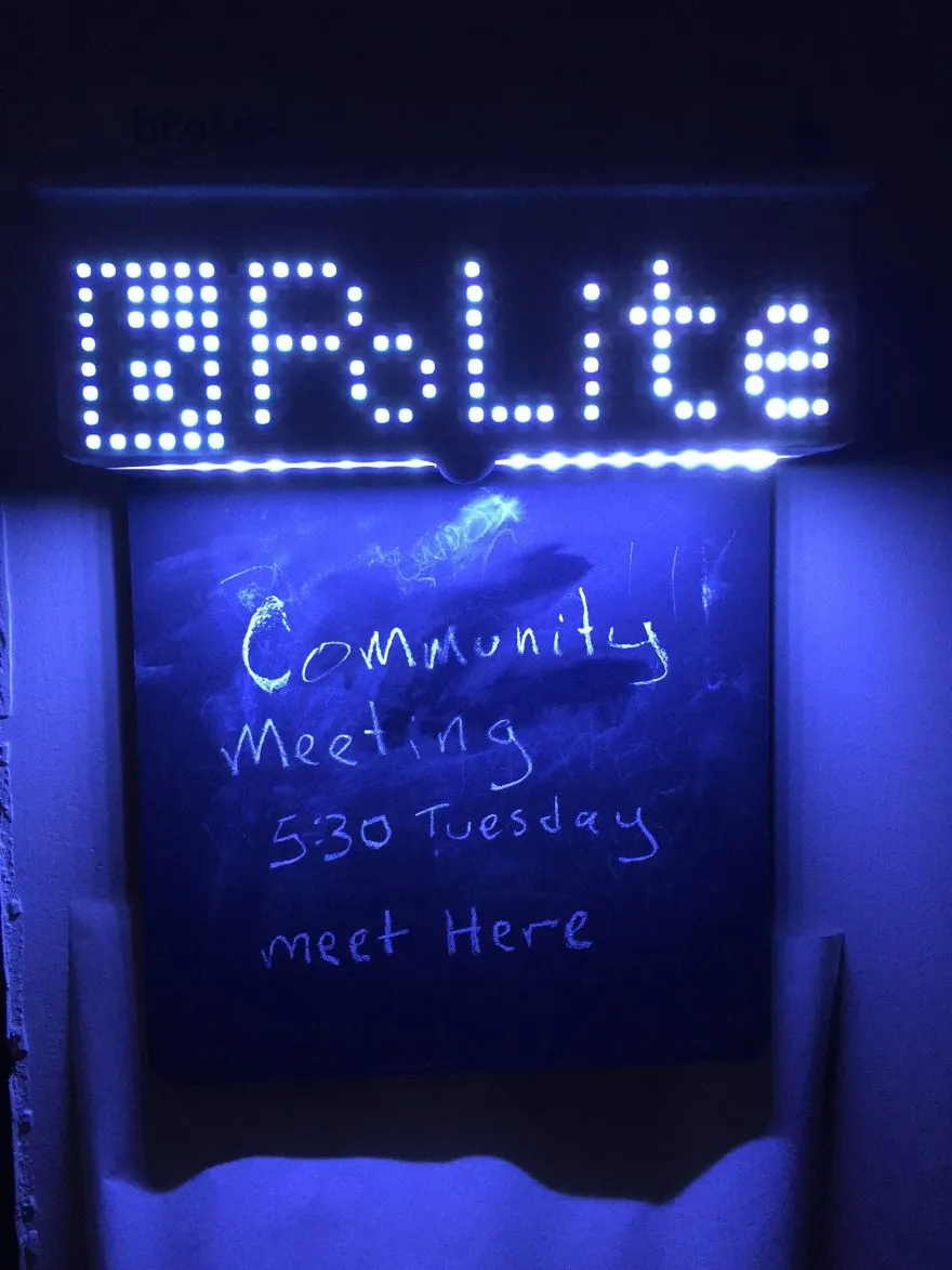 Photograph showing illuminated 'bPolite' board with dot-led display on the top and "community meeting 5:30 tuesday meet here" written on the chalkboard.