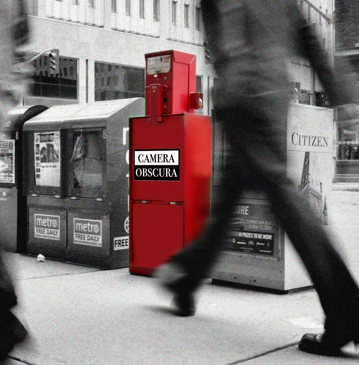 Black and white photograph of a sidewalk with a newspaper dispensing box in red labeled "Camera Obscura".