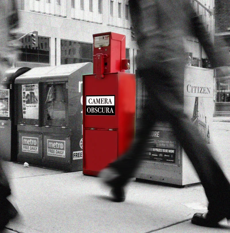 Black and white photograph of a sidewalk with a newspaper dispensing box in red labeled "Camera Obscura".