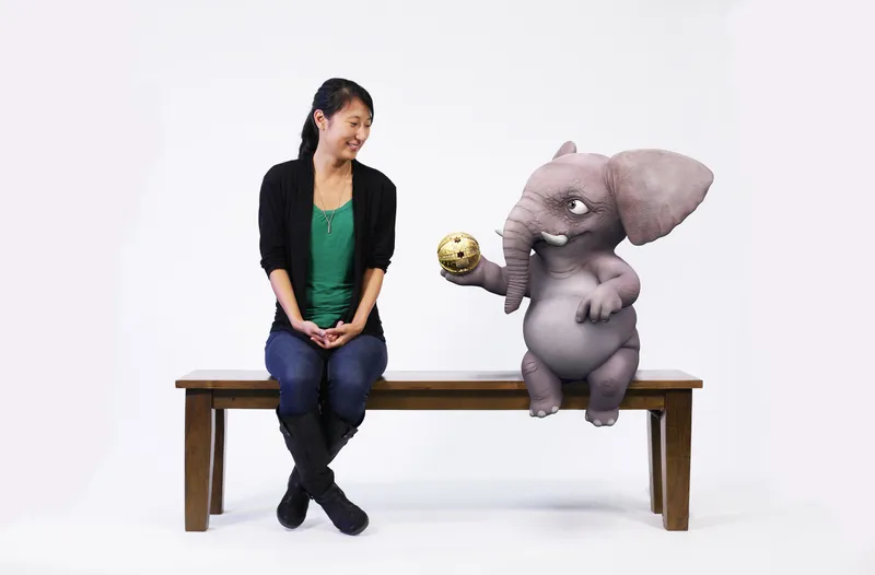 Image of a person and animated elephant sitting on a wood bench against a white backdrop. The elephant is looking at the person and handing over a golden orb.