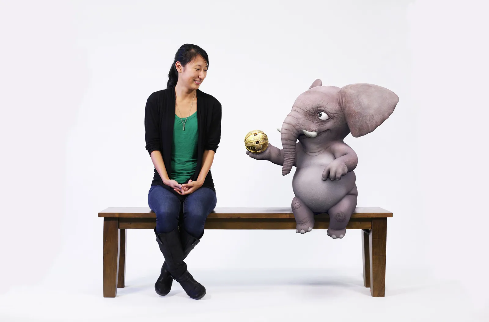 Image of a person and animated elephant sitting on a wood bench against a white backdrop. The elephant is looking at the person and handing over a golden orb.