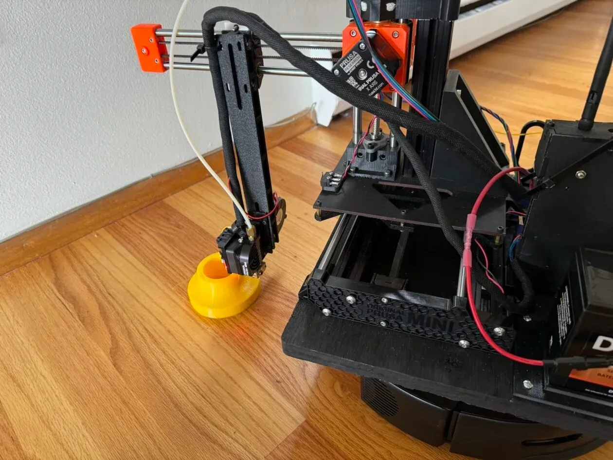 Photo of the 3D printer mounted on the robot vacuum as it is printing a yellow cylinder on a wooden floor.