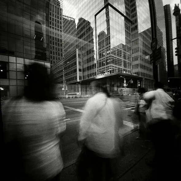 Black and white photo showing a sidewalk. Pedestrians and cars are motion-blurred and the buildings are clear.