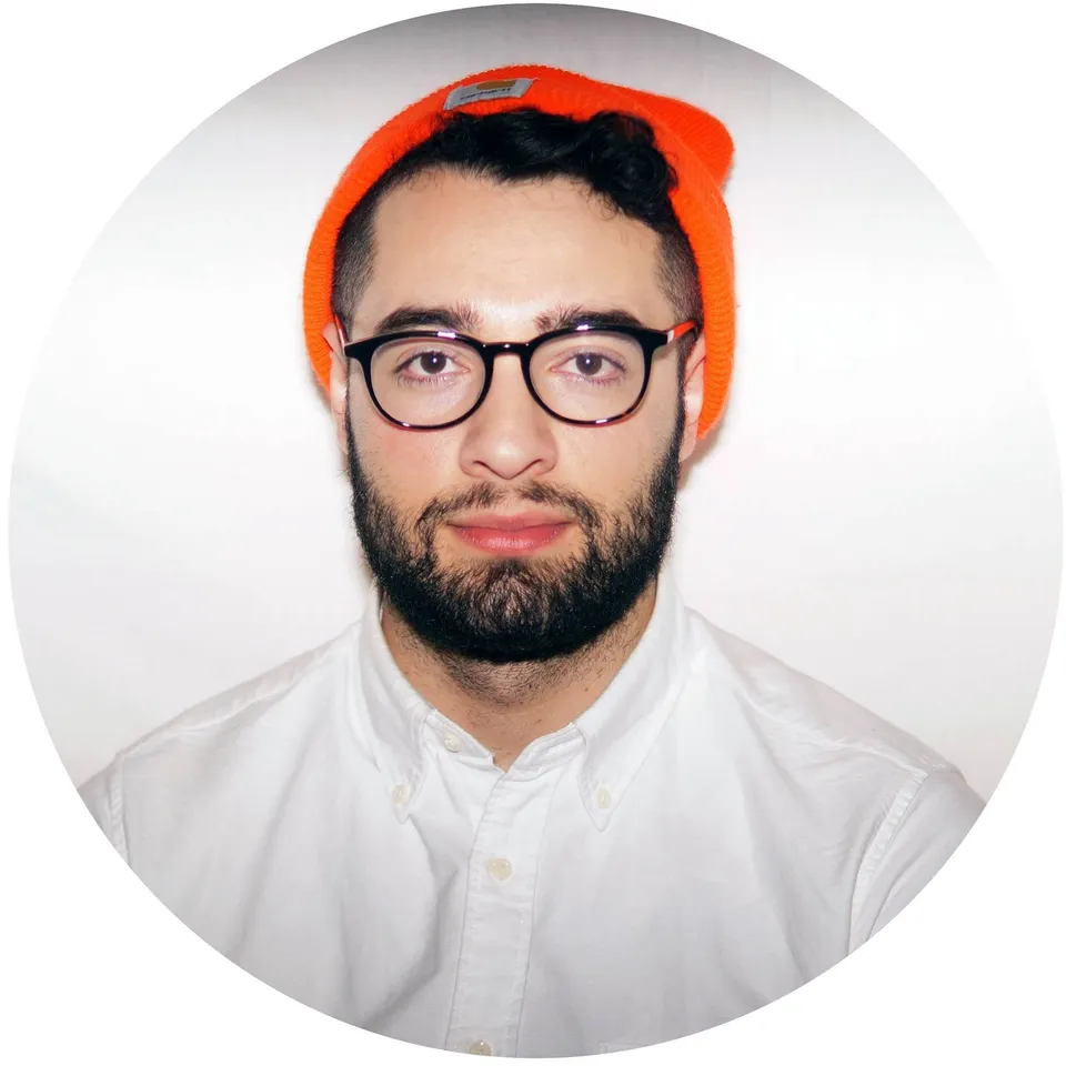 Portrait photo of a man wearing an orange beanie, white button up shirt, and dark glasses against a white background.