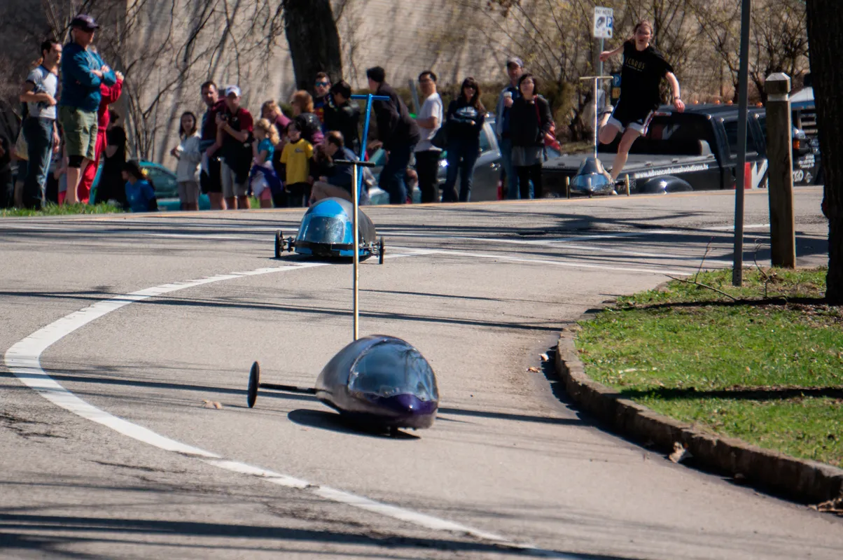 Two missile-shaped vehicles racing down a street as a crowd stands behind them.